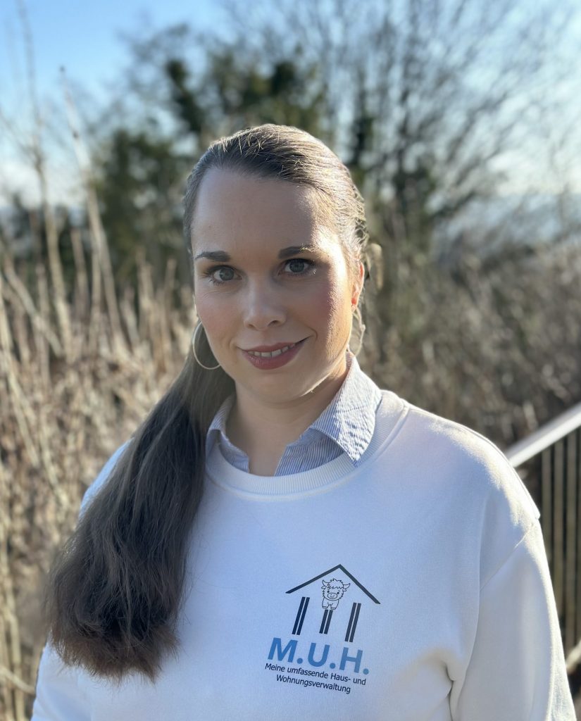 Foto von einer Frau mit braunem Pferdeschwanz, Hoop-Ohrringen und weißem Sweatshirt mit blauem M.U.H.-Logo im Sonnenlicht.
